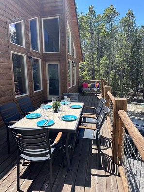 A wooden deck with a table set for dining, surrounded by chairs, and overlooking a forested area. - New! Spacious Bobcat lodge w/Hot Tub, Starlink (Fairplay)