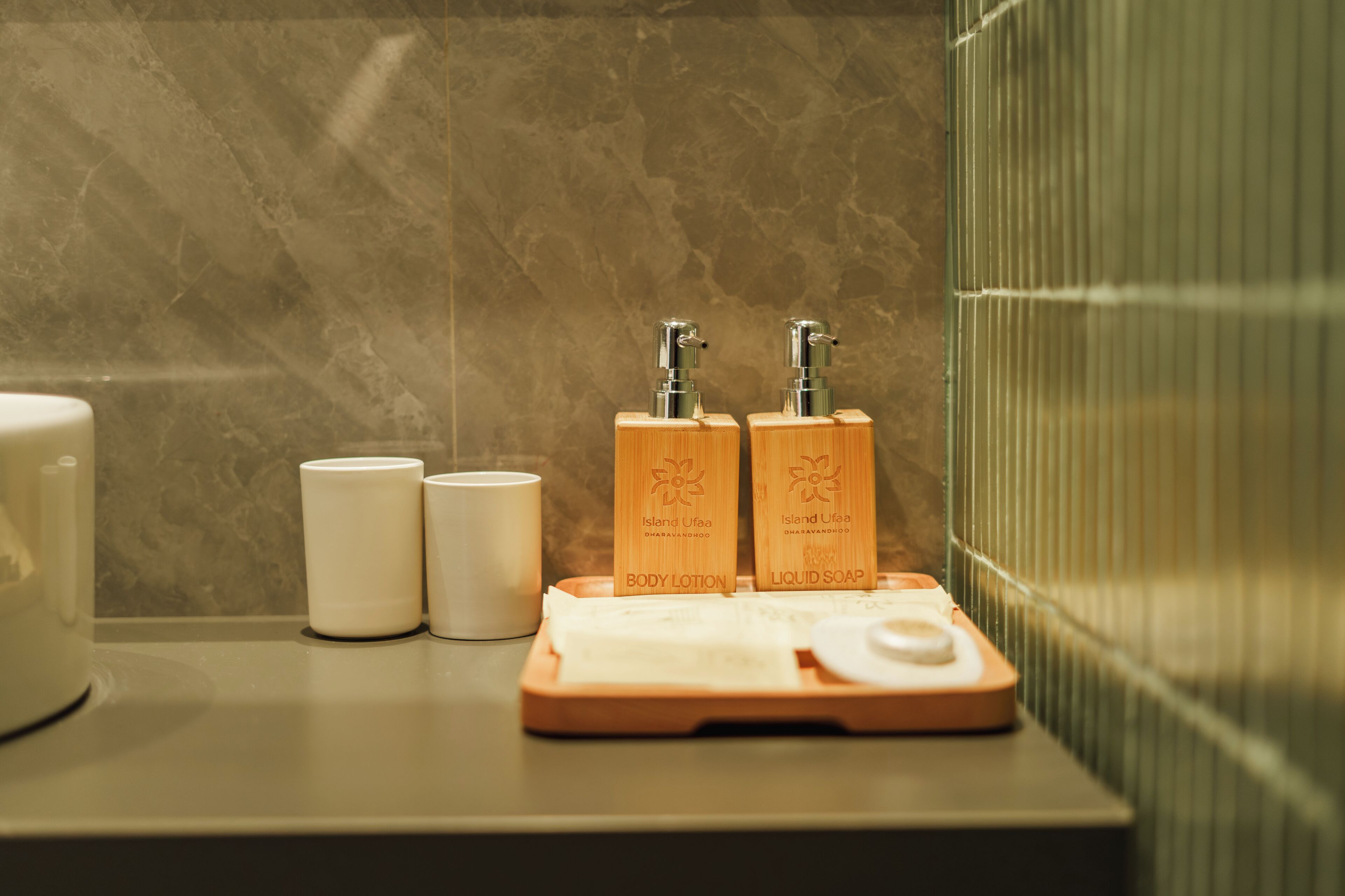 A bathroom with a marble wall, two wooden soap dispensers labeled 'Island Uffo,' a white soap dish, and a white cup.