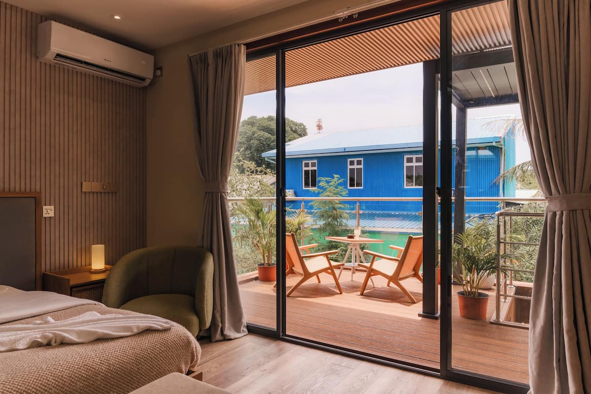 Habitación con cama, una silla, una mesa y balcón con vistas a una casa azul y vegetación.