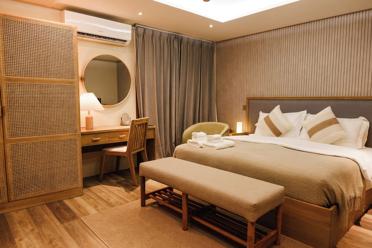 Habitación de hotel con cama, escritorio, silla, banco, espejo y ventana con cortinas.