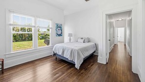 Ein Schlafzimmer mit Bett, großen Fenstern, Blick ins Grüne, einem Flur und einem Deckenventilator.