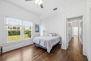 Ein Schlafzimmer mit Bett, großen Fenstern, Blick ins Grüne, einem Flur und einem Deckenventilator.