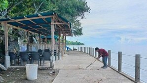 Ein Betonweg mit einer Holzkonstruktion und Stühlen links, eine Person fegt in der Nähe des Wassers.