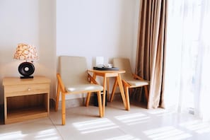 Una habitación moderna con una mesita de noche de madera, una lámpara, dos sillas, una pequeña mesa y una ventana con cortinas.