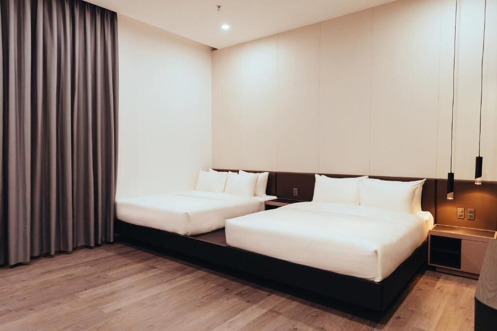 Quarto de hotel moderno com duas camas, piso de madeira, uma janela grande com cortinas e uma mesa de cabeceira com abajur.