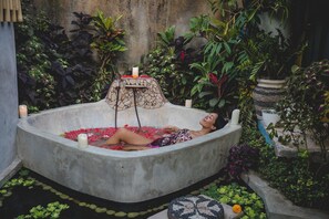 Eine Person entspannt sich in einer Betonbadewanne, gefüllt mit Wasser und Rosenblättern, umgeben von üppigem Grün und Topfpflanzen.