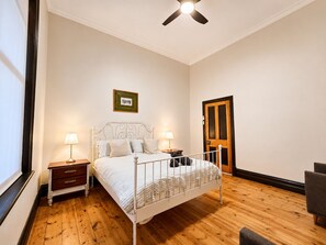 A bedroom with a bed, bedside tables, a ceiling fan, a wooden door, and a framed picture on the wall.