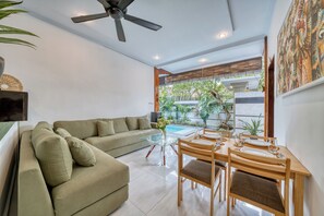 Ein modernes Wohnzimmer mit einer Couch, Esstisch, Stühlen, Deckenventilator und Blick auf einen Swimmingpool und Grünflächen.