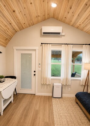 Ein Zimmer mit Holzboden, einer weißen Tür, einem Fenster mit Vorhängen, einem kleinen Tisch mit Pflanze, einem Bett und einem Heizkörper.