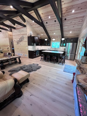 Un vaste espace de vie doté d’un grand plafond en bois, d’un îlot central avec des tabourets, de meubles de rangement en bois foncé et d’une vue sur la cuisine.