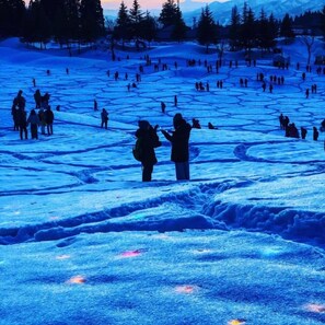 Un vaste espace extérieur où l’on voit des gens se promener et rester debout, entouré d’arbres et avec une chaîne de montagnes en arrière-plan.