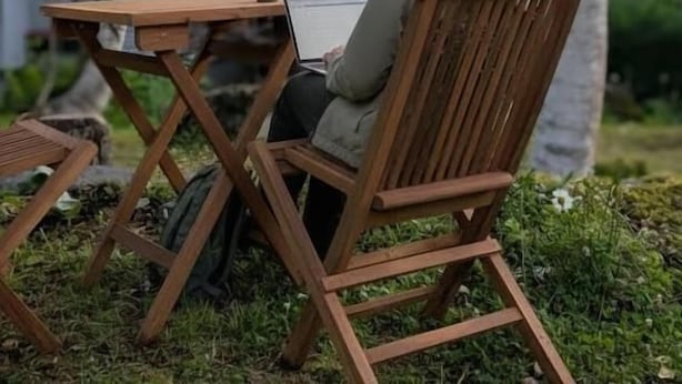 Une personne est assise à une table en bois à l’extérieur, devant un ordinateur portable, et est entourée de verdure et d’arbres.