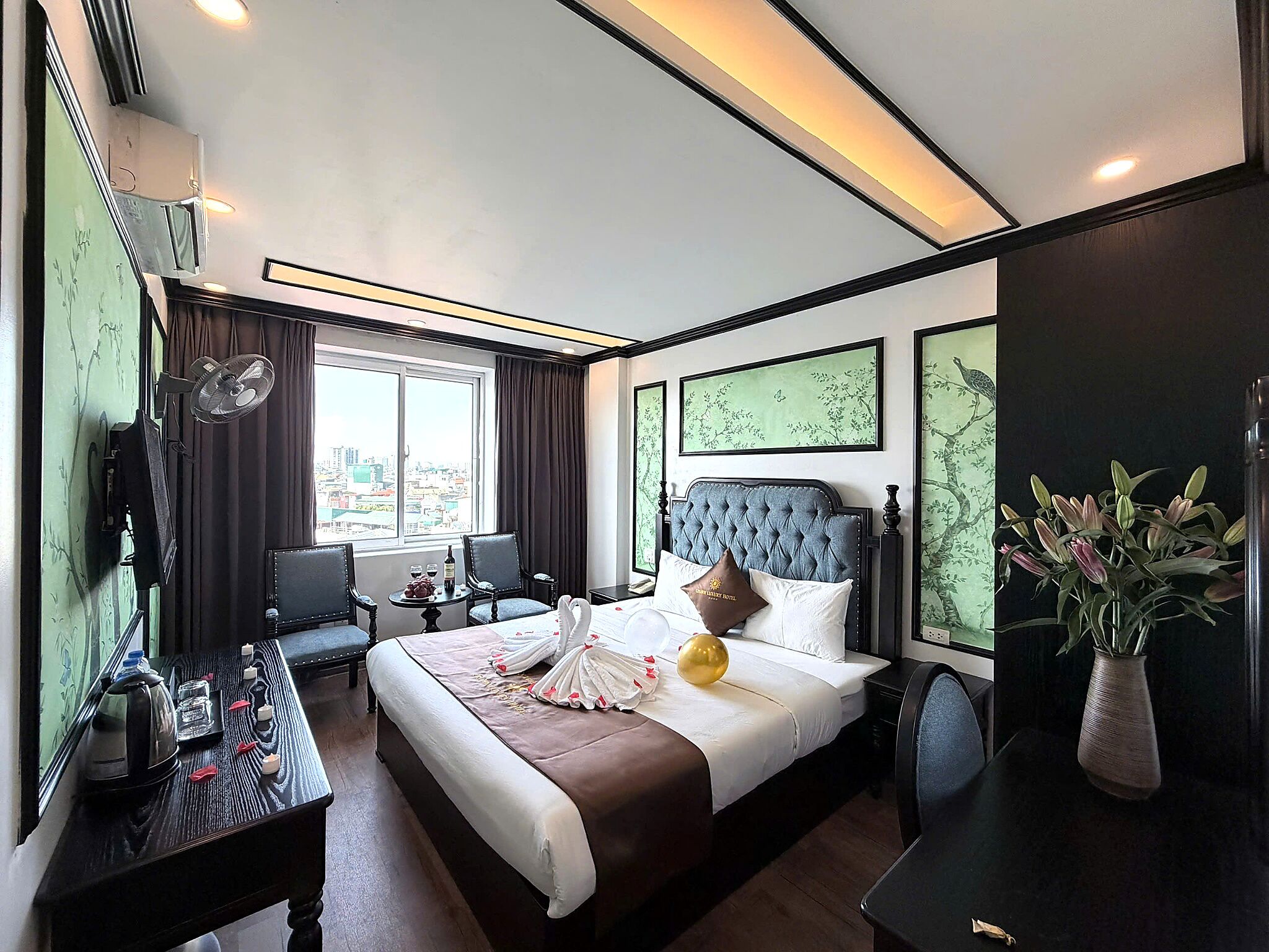 Una habitación de hotel con una cama grande, una zona de estar con una mesa y sillas, vistas de la ciudad a través de la ventana y obras de arte en las paredes.