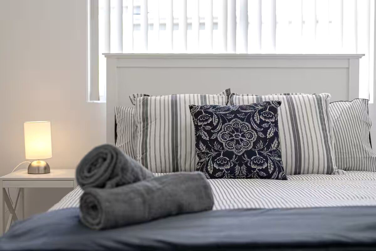 A neatly made bed with striped bedding and a decorative pillow, a bedside lamp, and a rolled towel on a white nightstand.