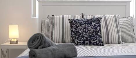 A neatly made bed with striped bedding and a decorative pillow, a bedside lamp, and a rolled towel on a white nightstand.