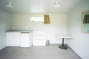 A compact room with a kitchenette, a small window with curtains, a white table, and a trash bin. - Welcome Bay Hot Pools and Accommodation (Tauranga)