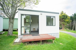 Una casa móvil moderna de un solo piso con terraza de madera, grandes puertas correderas de vidrio y un interior visible que incluye una cama y una pequeña mesa.