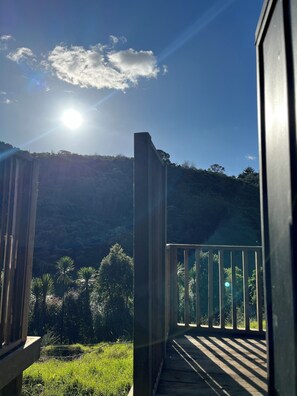 A balcony with a view of a forested hillside, a clear sky, and the sun shining. - Welcome Bay Hot Pools and Accommodation (Tauranga)