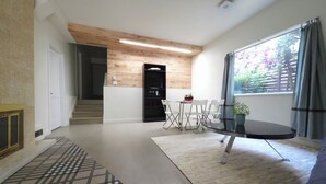 A modern living room with a glass coffee table, a dining area with a small table and chairs, a staircase leading to an upper floor, and a large window with curtains. - Private Creekside Garden Suite - Patio, Deck, BBQ & Free Parking. Pet Friendly. (North Vancouver)