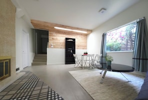 Ein modernes Wohnzimmer mit Glastisch, ein Essbereich mit kleinem Tisch und Stühlen, eine Treppe, die zu einer oberen Etage führt, und ein großes Fenster mit Vorhängen.
