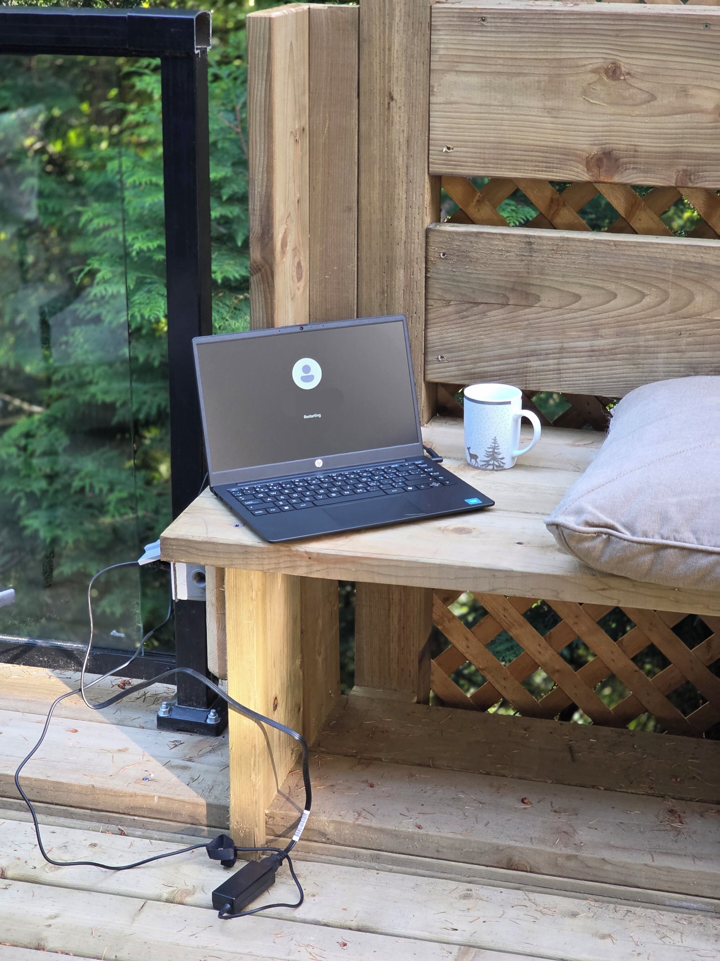 A laptop on a wooden table with a cup and a pillow, connected to a power source.