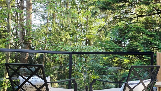 A deck with two chairs, a round table with a drink, and a view of a forest. - Private Creekside Garden Suite - Patio, Deck, BBQ & Free Parking. Pet Friendly. (North Vancouver)