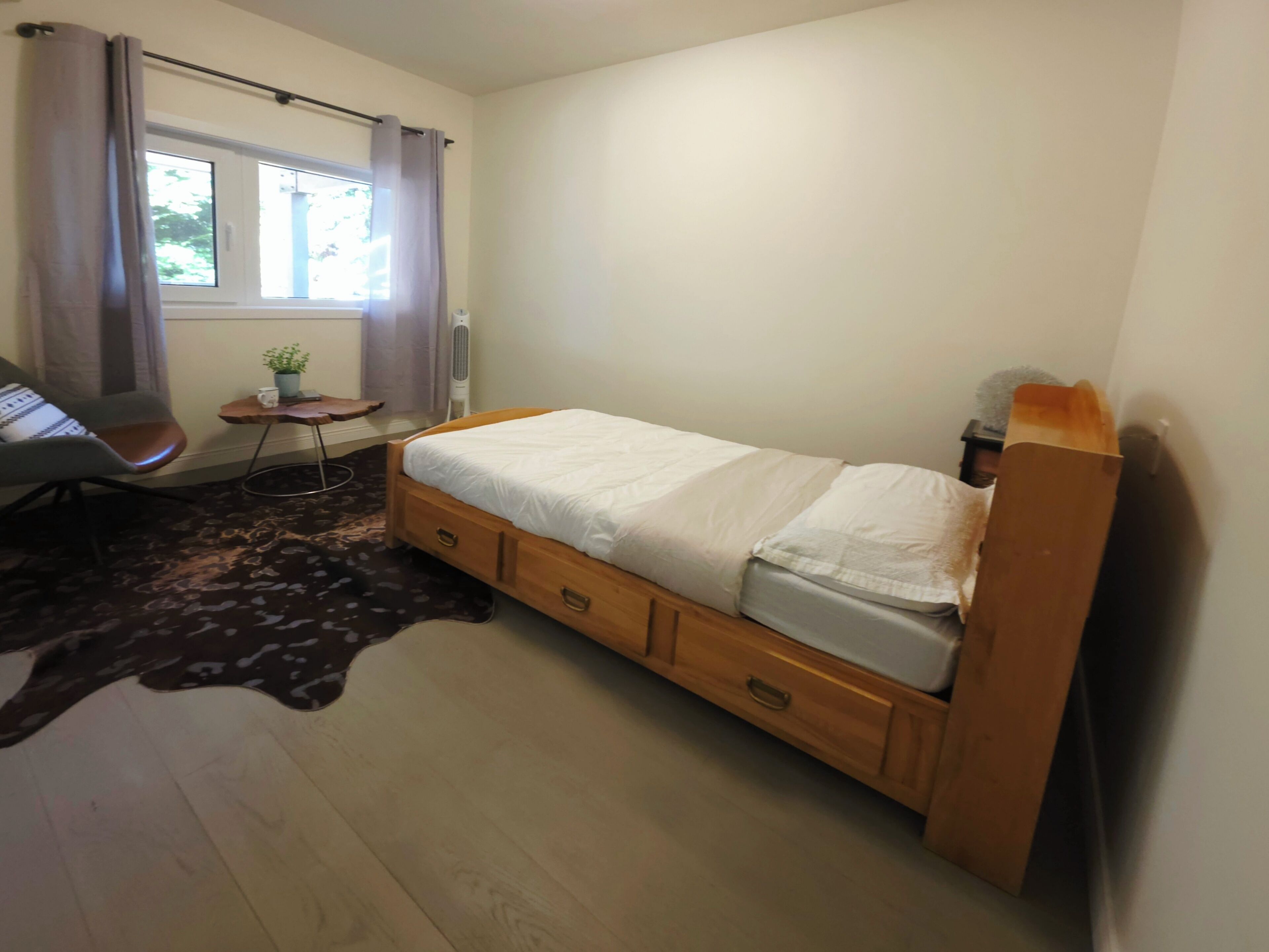 A bedroom with a bed, a chair, a small table with a plant, and a window with curtains.