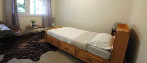 A bedroom with a bed, a chair, a small table with a plant, and a window with curtains.