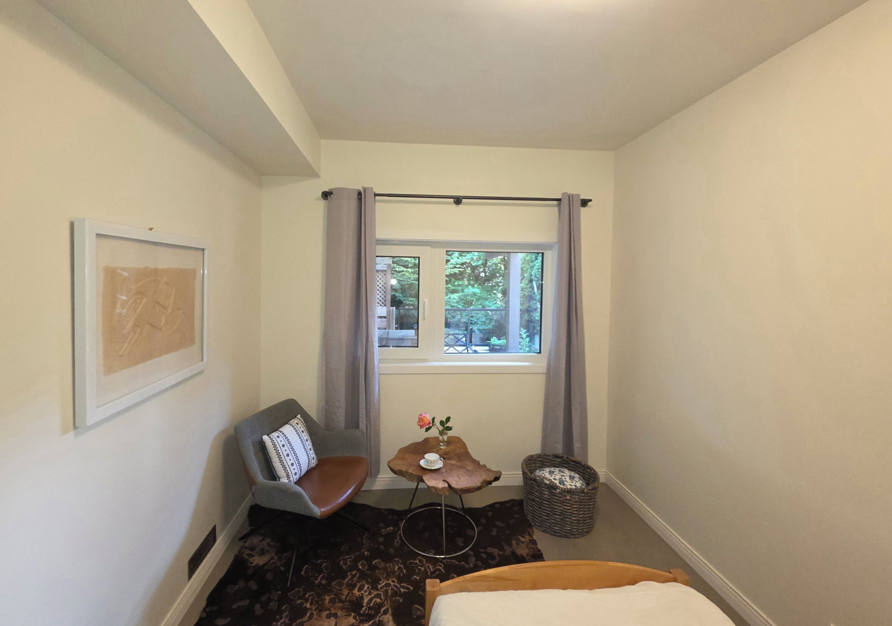 A small hotel room with a bed, a chair, a small table with a vase, a window with curtains, and a framed picture on the wall.