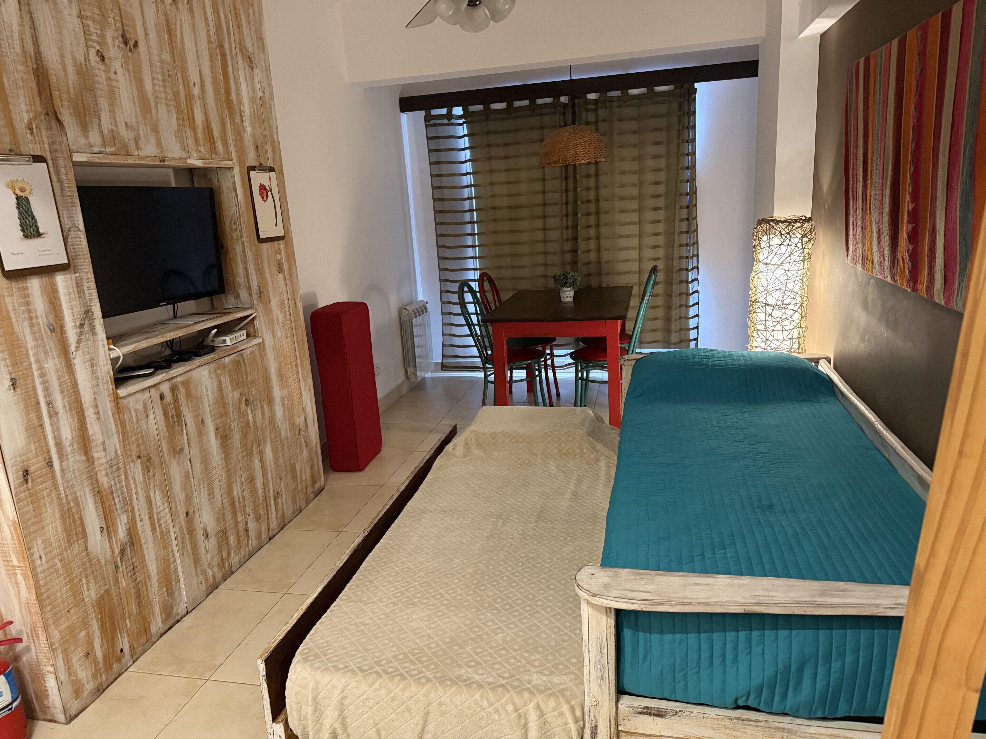 Quarto com parede de madeira, televisão, um banquinho vermelho, cama verde-água, mesa de jantar com cadeiras e janela com cortinas listradas.