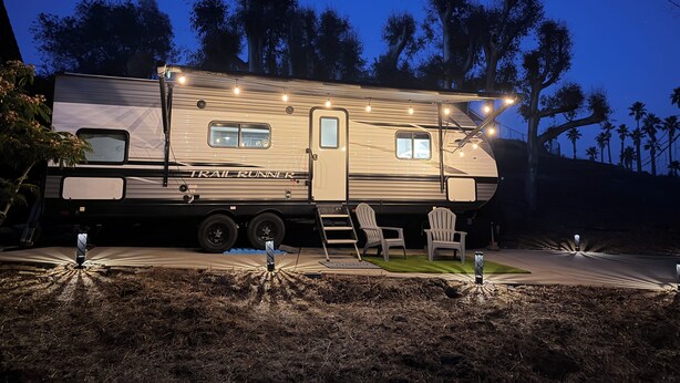 Een grote camper met een 'TRAIL RUNNER' bord, een overdekte zitplaats buiten, en een goed verlichte oprit.