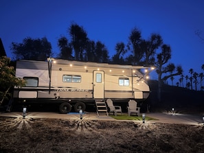 A large RV with a 'TRAIL RUNNER' sign, a covered outdoor seating area, and a well-lit driveway.