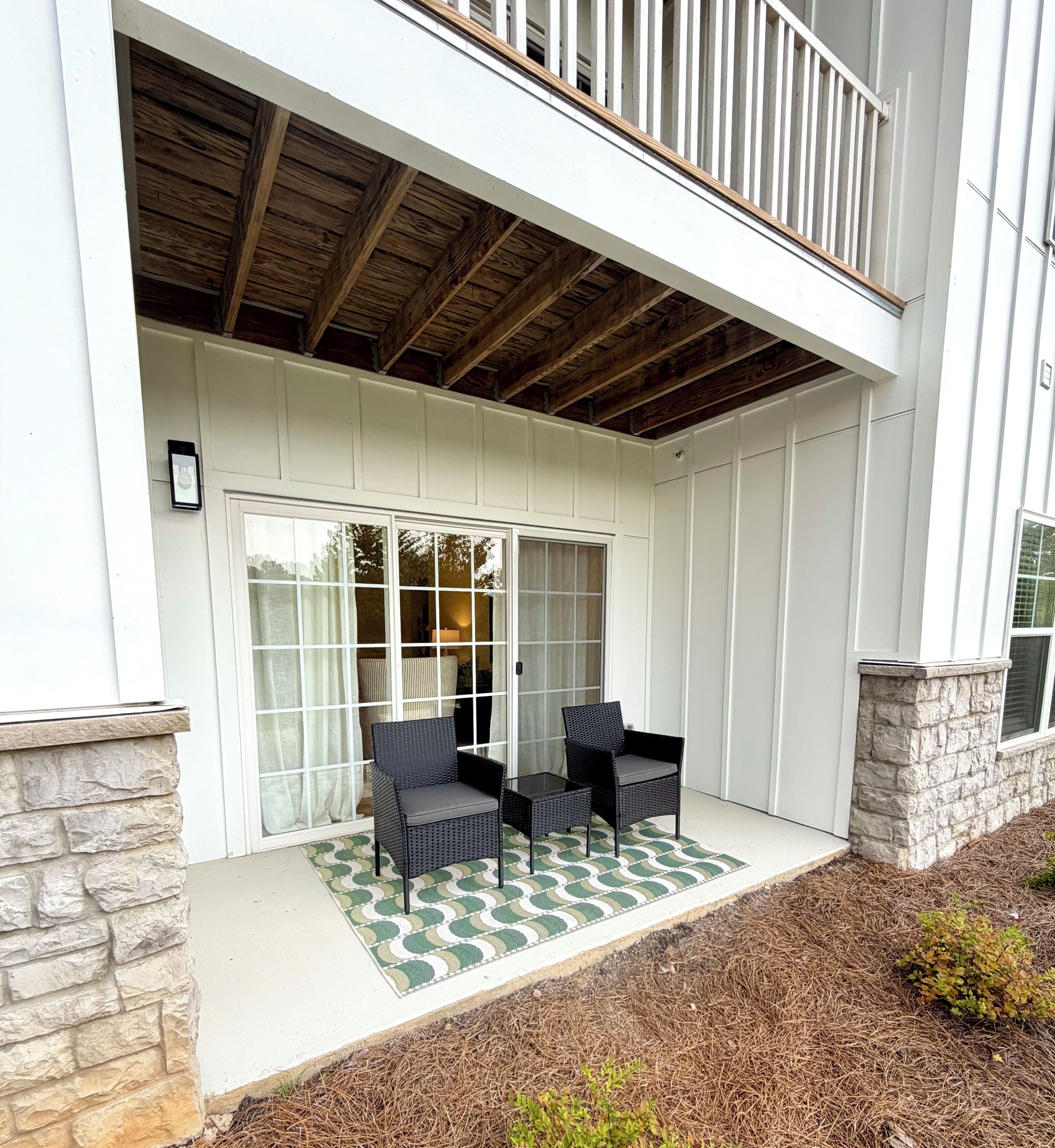 A covered patio with two chairs and a table, a stone wall, and a wooden ceiling.