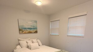 A bedroom with a bed, a painting, two windows with blinds, and a ceiling light.