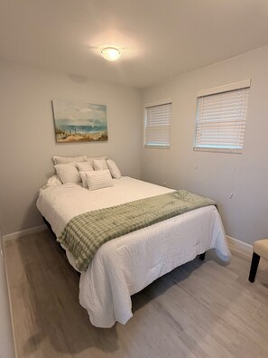 Ein Schlafzimmer mit einem Bett, einem Gemälde, zwei Fenstern mit Jalousien und einer Deckenleuchte.