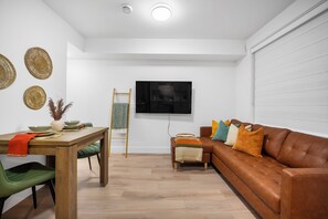 A modern living room with a wooden dining table, green chairs, a brown leather sofa, and a flat-screen TV mounted on the wall.