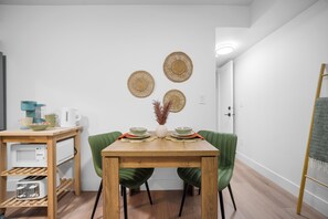 Ein Essbereich mit einem Holztisch, zwei grünen Stühlen, einer weißen Vase mit Trockenpflanzen und drei gewebten Wanddekorationen.