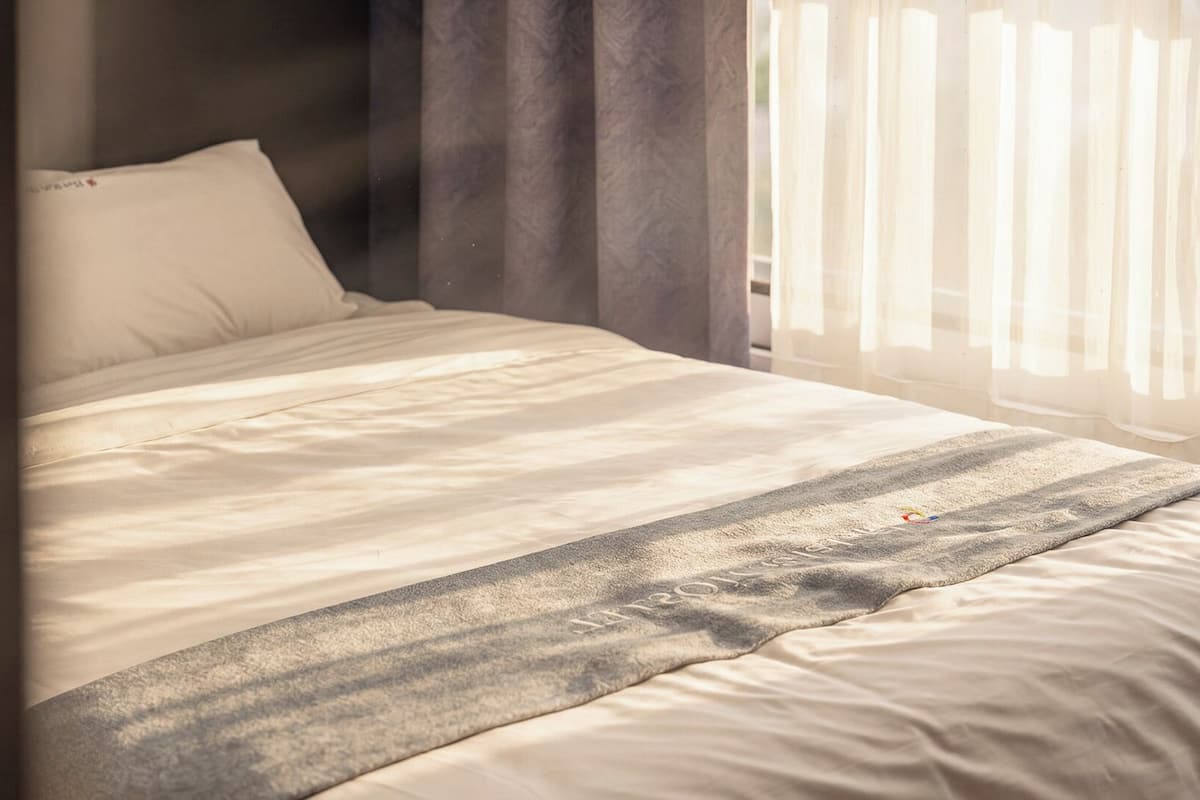 Una cama con edredón blanco y manta gris, una almohada y una ventana con cortinas traslúcidas.