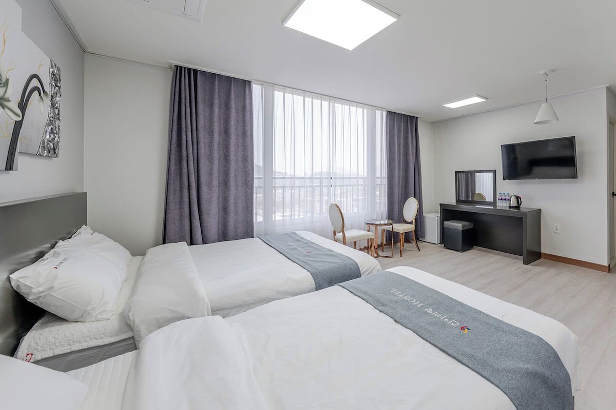 Habitación de hotel con dos camas, un escritorio, una silla, un televisor y un ventanal con cortinas.