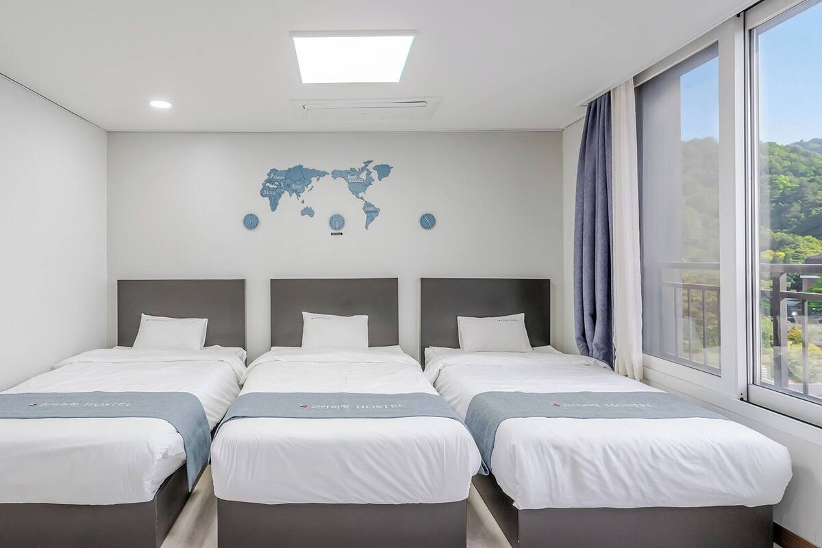 Habitación de hotel con tres camas, un mapa mundial en la pared y una ventana con vistas a zonas verdes.