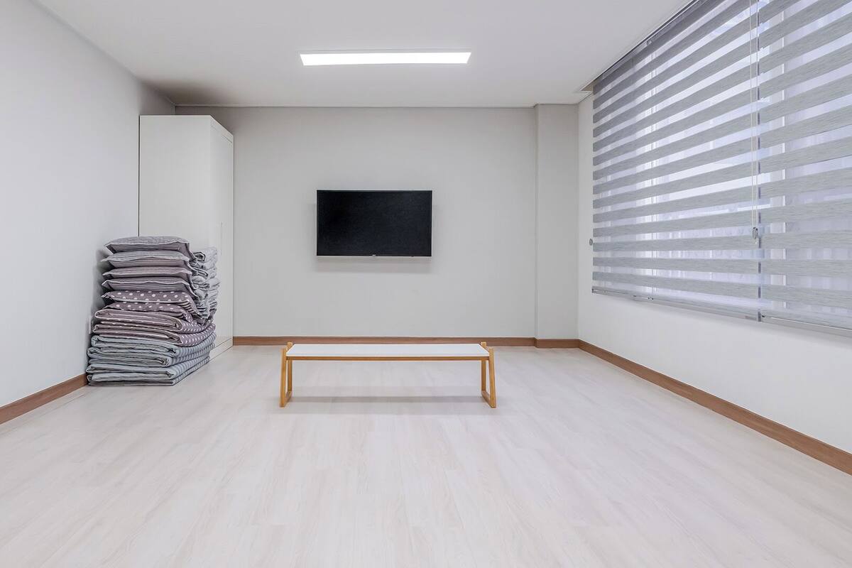 Una habitación con un banco de madera, un montón de mantas dobladas, un cuadro con marco negro en la pared y una ventana con persianas.
