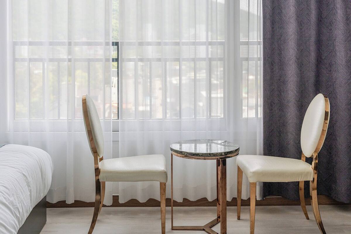 Una habitación de hotel con una cama, dos sillas, una mesita y una ventana con cortinas ligeras.