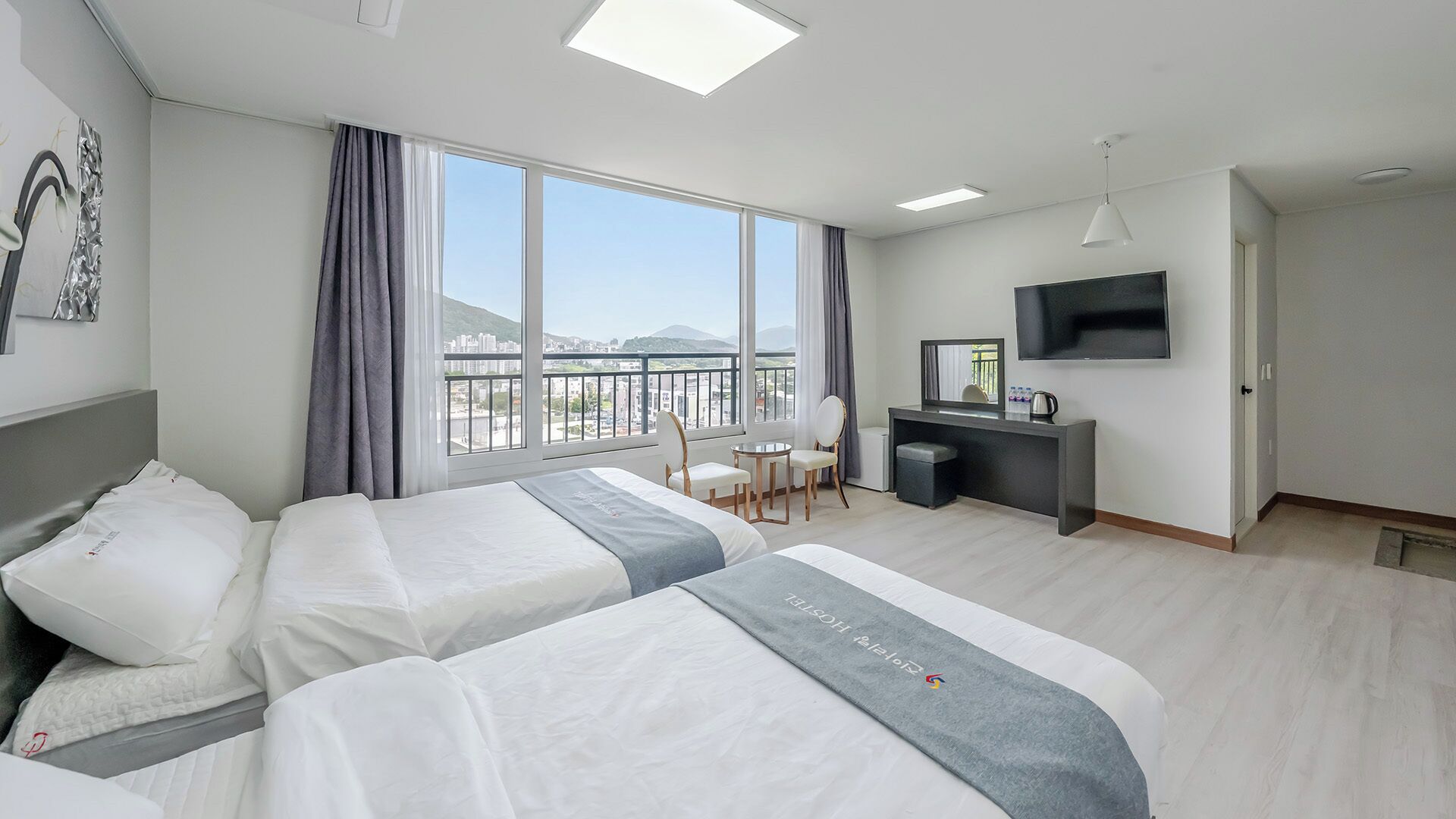 Quarto de hotel com duas camas, uma escrivaninha, uma cadeira, televisão e uma janela grande com vista.