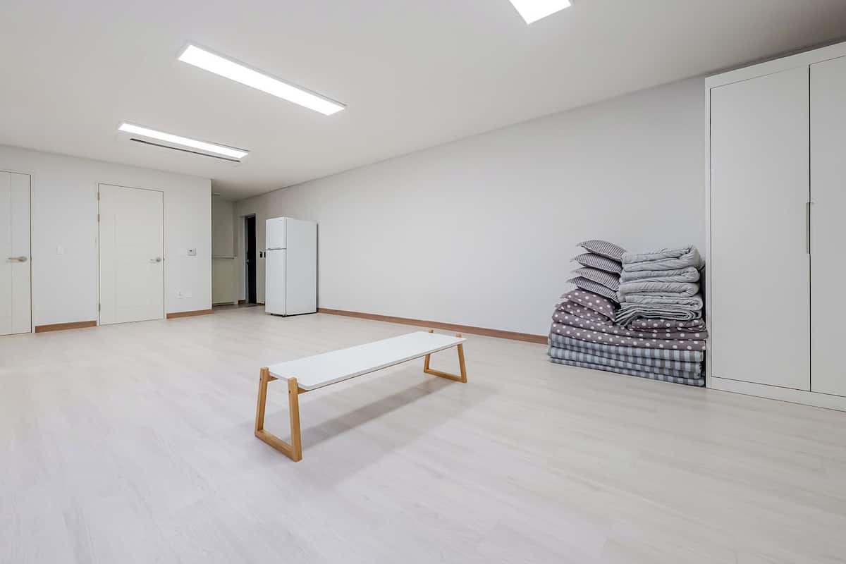 Una habitación amplia con un largo banco, un refrigerador y ropa de cama apilada ordenadamente.