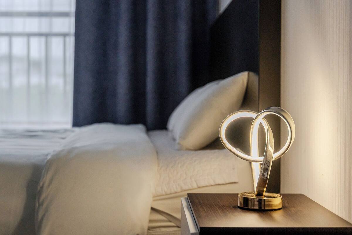 Habitación de hotel con cama, mesita de noche y un objeto decorativo, además de una ventana con cortinas.