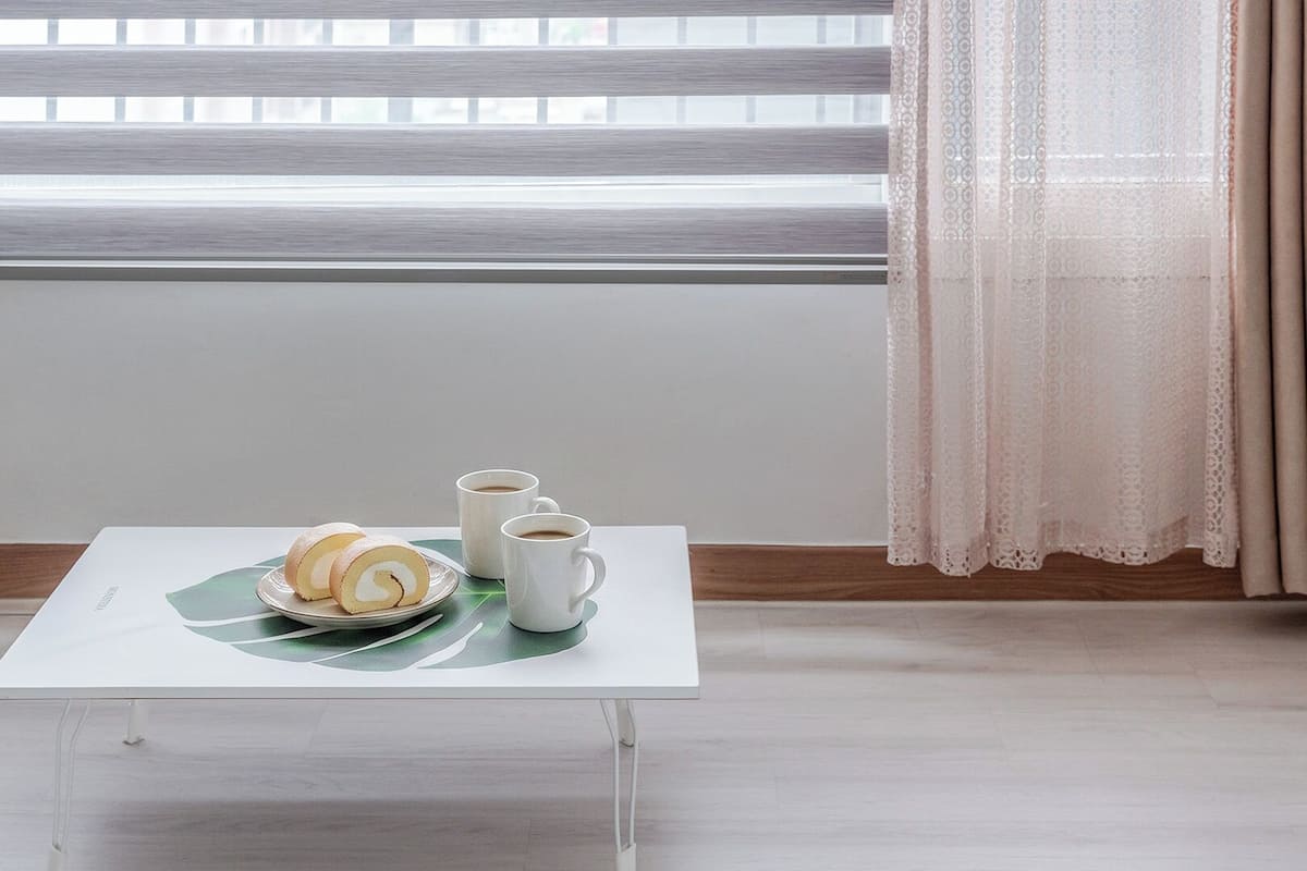 Una pequeña mesa con un plato de pasteles y dos tazas de café, una ventana con persianas y una cortina.