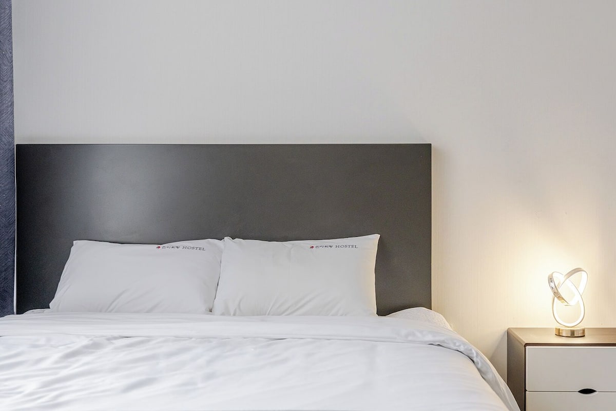 Una habitación de hotel con cama, almohadas, mesita de noche y una lámpara en la pared.