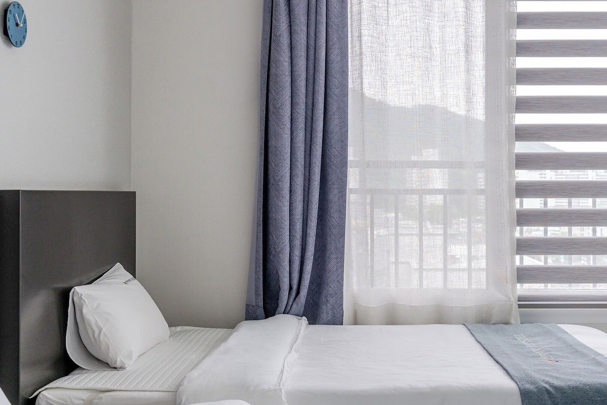 Habitación de hotel con cama, cabecera, ventana con persianas y un reloj en la pared.