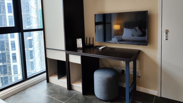 Une chambre d’hôtel moderne dotée d’une grande fenêtre, d’un téléviseur à écran plat fixé au mur, d’un bureau avec une chaise, d’un petit pouf et d’une étagère intégrée au-dessus du bureau.