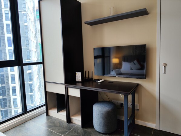 A modern hotel room with a large window, a flat-screen TV mounted on the wall, a desk with a chair, a small ottoman, and a built-in shelf above the desk. - Portrait Hotel (Petaling Jaya)